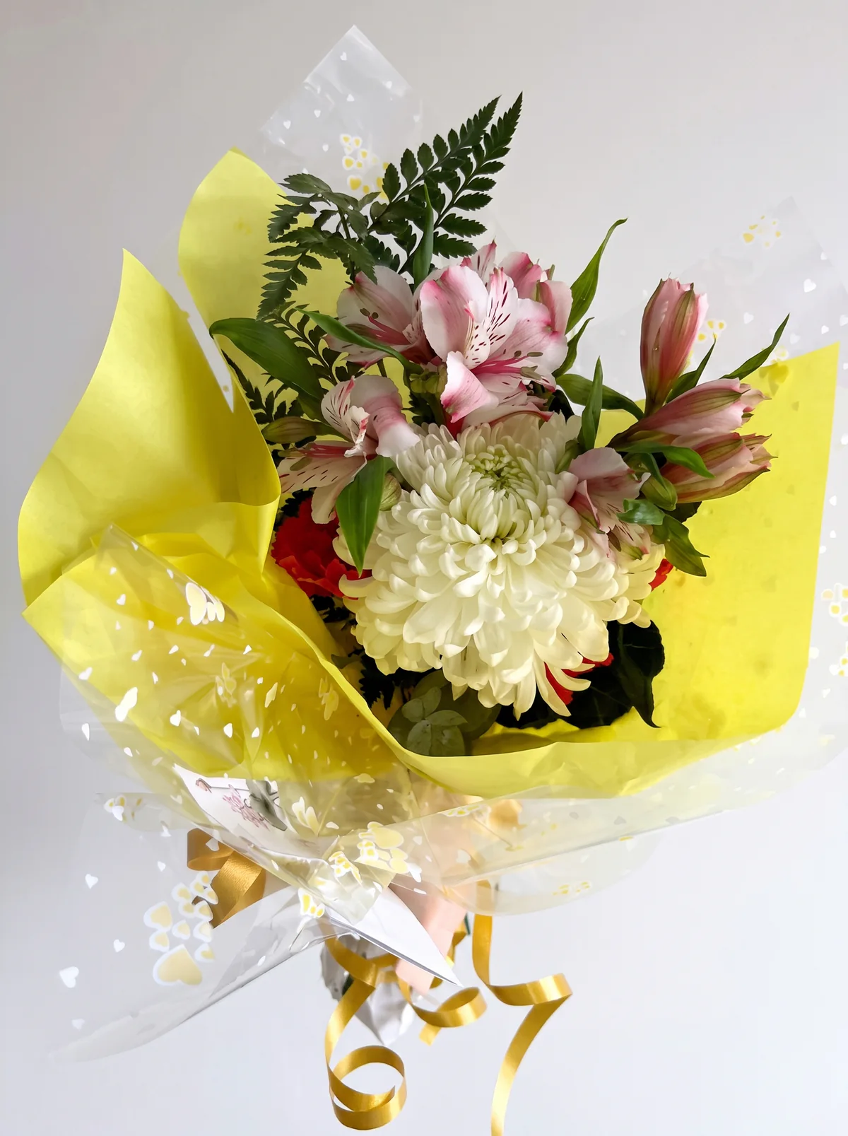 Ramo con una flor blanca de pétalos abundantes, flores rosadas y rojas y follaje, con envoltorio amarillo y cinta dorada.