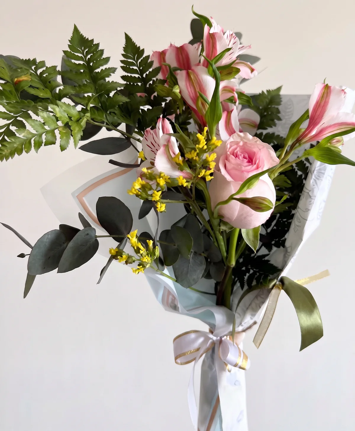 Ramo de flores rosadas, pequeños racimos amarillos y follaje, con envoltorio claro y lazos blanco y verde oliva.