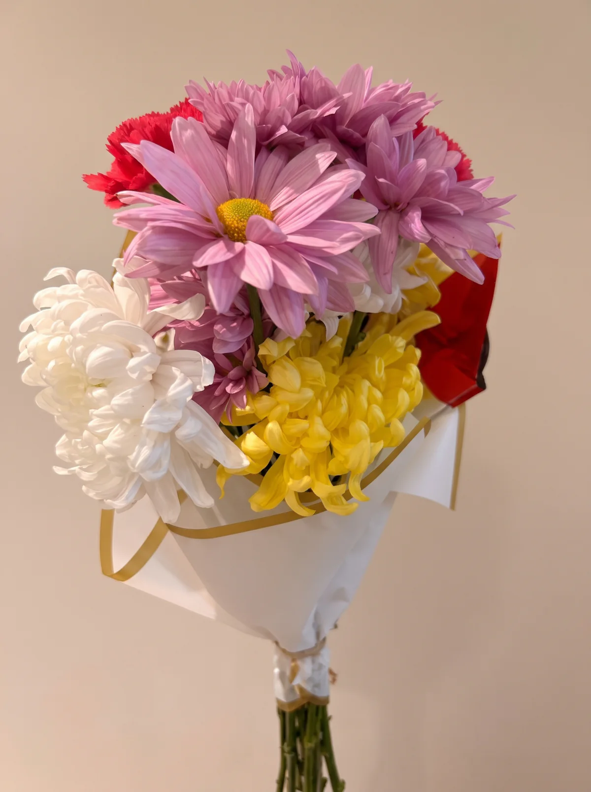 Ramo de flores lilas, blancas, amarillas y rojas, con envoltorio blanco de borde dorado y tallos a la vista.